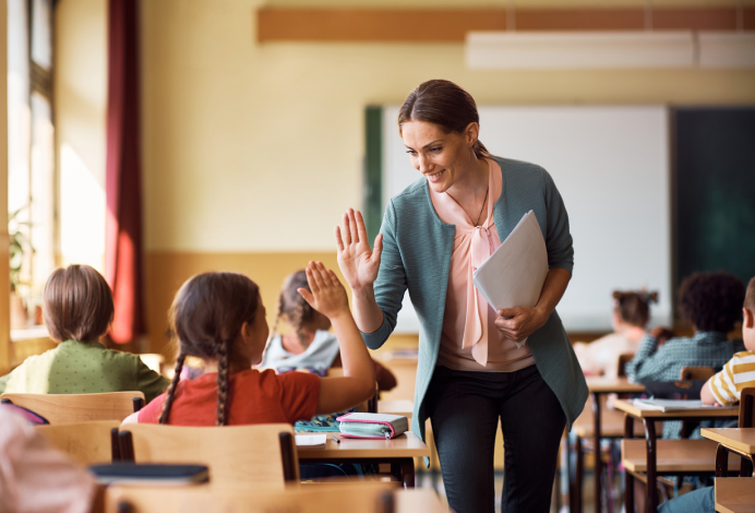 A woman holds a folder, standing in front of her attentive students, engaged in a classroom setting.